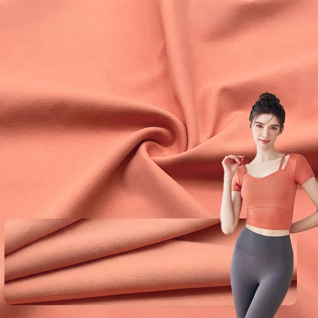 A product showcase image featuring a large section of draped, textured coral pink fabric. The fabric has a soft, suede-like, double-sided brushed appearance. In the lower-left corner, a detail inset shows stacked layers of the same fabric. On the right, a smiling model stands, wearing a matching coral pink short-sleeved crop top and grey high-waisted leggings, demonstrating the fabric's look on a finished garment. The overall aesthetic is clean and high-quality, set against a light background with subtle Chinese text.