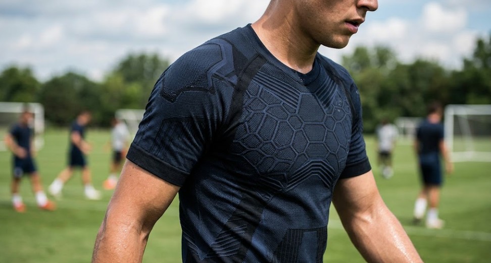 A close-up, authentic photograph of a male soccer athlete on a green field during a workout. The focused area on his navy geometric knit technical sports jersey shows intricate cellular and interlocking patterns designed for sweat vapor management. His neck and arms are glistening with sweat, illustrating active field conditions.