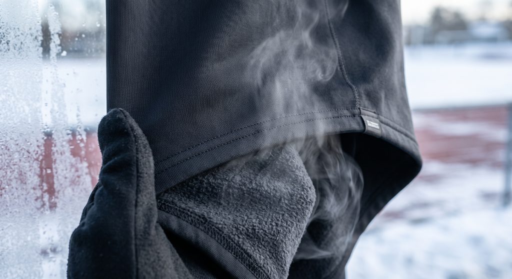 A macro studio photograph showcasing the stretch and breathability of performance fleece sports fabric. A gloved hand slightly pulls the dark gray fabric, which reveals a soft brushed underside and a smooth outer layer. Subtle wisps of vapor are seen rising through the fabric to illustrate moisture escape, set against a blurred frosty background suggesting freezing winter conditions.