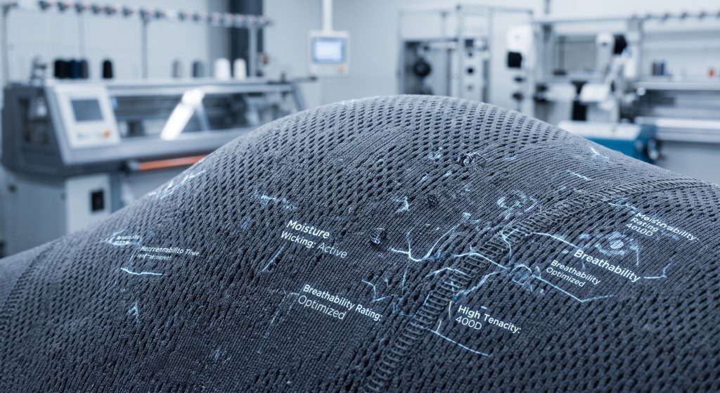 Macro close-up of dark slate-gray pro sports fabric showing breathable geometric knit pattern and integrated technical data visualization overlay for wicking and durability.