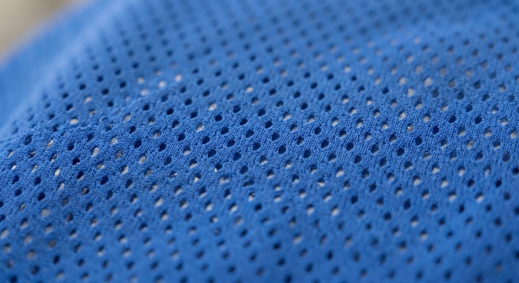 A photorealistic macro shot of cobalt blue synthetic birdseye mesh fabric with engineered geometric micro-perforations, used for lightweight and breathable basketball jerseys.