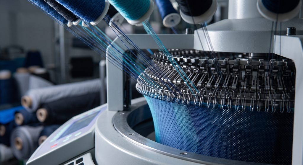 A close-up photograph of a circular knitting machine actively forming a flexible, breathable interlock fabric by interlocking synthetic filaments for high-performance activewear.