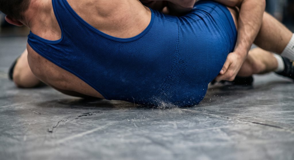 A raw, textured close-up photograph showing the dense interlocking knit pattern of a professional wrestling singlet panel resisting abrasion as it is dragged forcefully across a gray, worn wrestling mat surface.