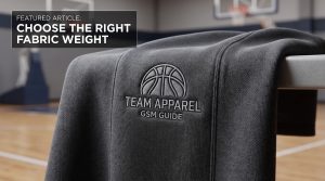 Detailed closeup of textured basketball fleece fabric with an embroidered basketball logo and "TEAM APPAREL GSM GUIDE" text, set against a blurred basketball court background.