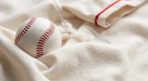 Macro photograph of soft, creamy white 100% cotton fabric weave with a traditional stitched baseball resting on it, highlighting its comfort and performance for team uniforms.